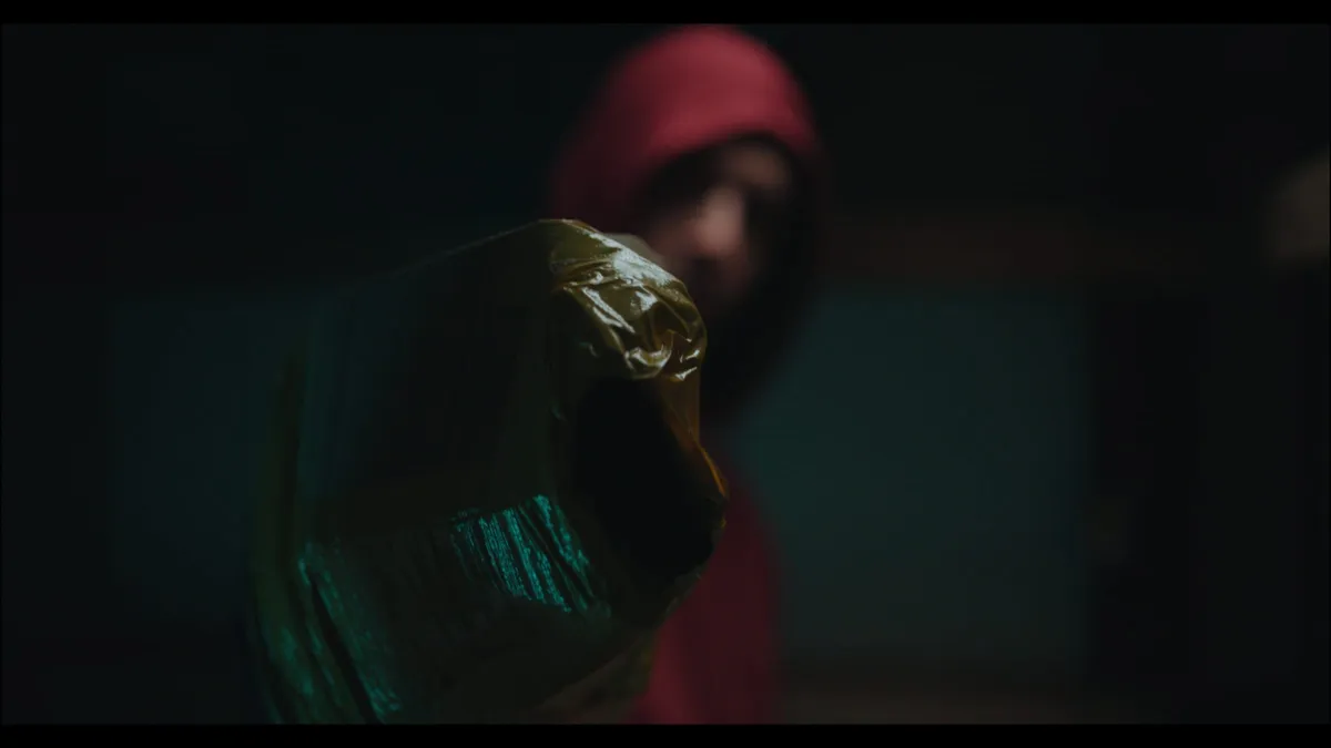 A dark, wrapped object held towards the camera in close focus, with a hooded figure in red blurred behind in dim light.