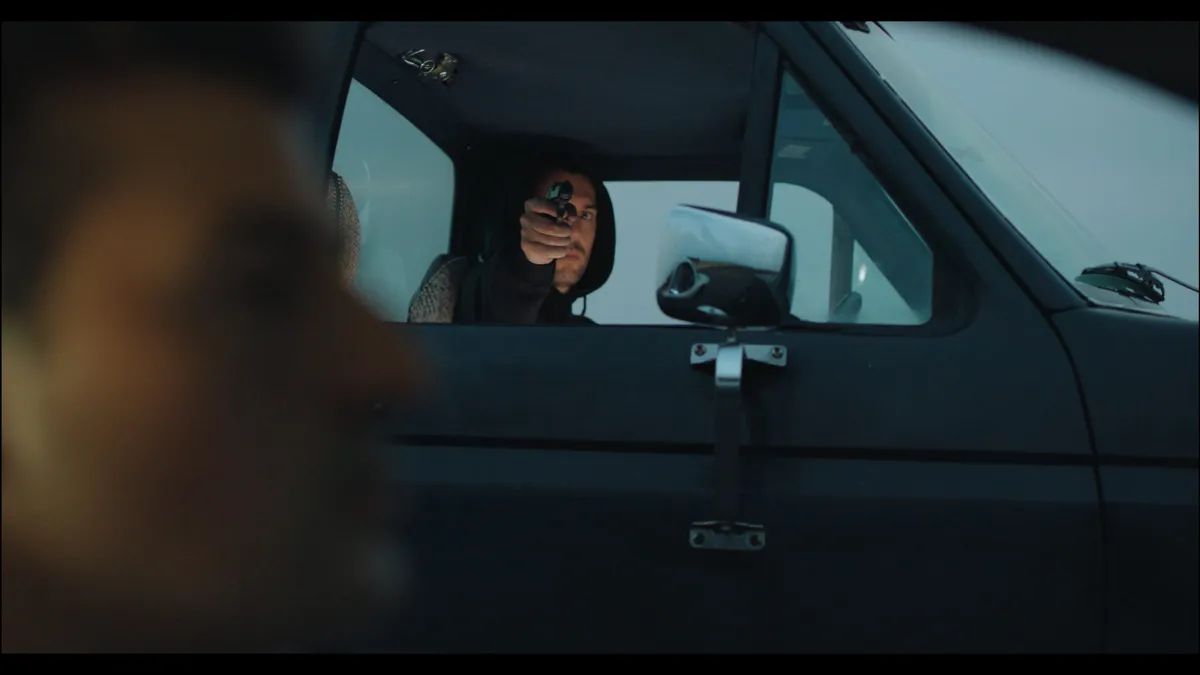 A hooded figure in a pickup points a handgun through the open window, seen past an out-of-focus figure in the foreground.