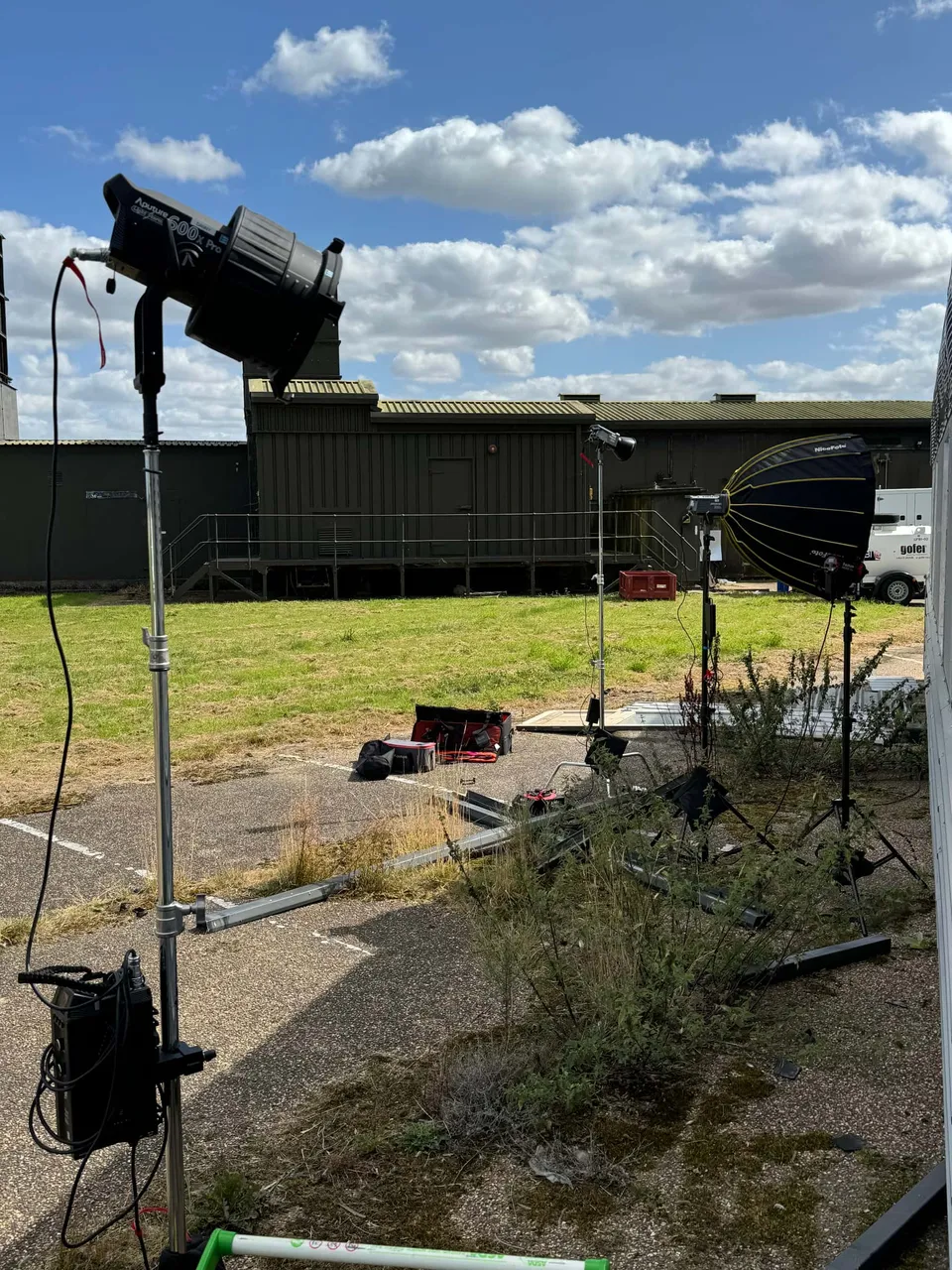 Lighting stands and a softbox rigged outdoors on tarmac beside a grass verge, with portacabins and a car park under a partly cloudy sky.